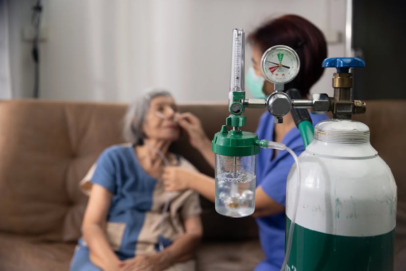 Elderly woman receiving oxygen therapy with a nasal cannula at home, assisted by a healthcare worker.