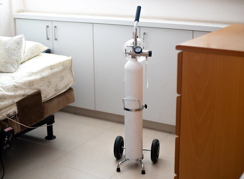 A portable oxygen cylinder with a regulator and wheels positioned beside a bed in a home setting.